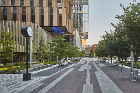 Brick walkway in the foreground with a clock sculpture on the left and a building on the right with two LED sculptures hanging from the buildings overhang.