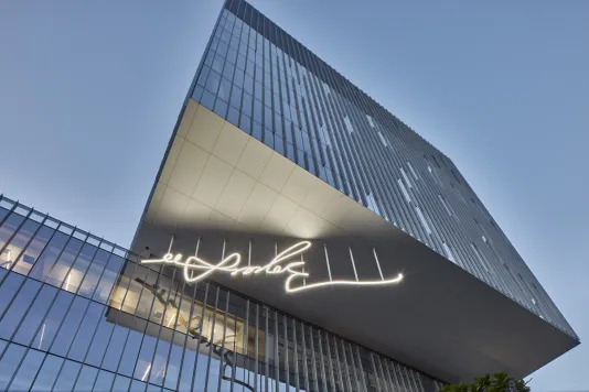 View from below of a glass building with an overhang that jets out and has a white LED sculpture of a signature hanging from it.