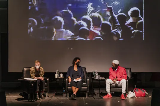 Three panelists for Altered Access sit on a stage engaged in a panel discussion. Behind them, is the lower third of a projector screen with a blurry image of a crowd projected.