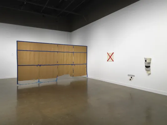 On the left, a large brown rectangular tapestry with blue gridded lines hangs from the ceiling. The tape-like lines divide the tapestry into three rows and four colors. On the right, three smaller fiber artworks are displayed. Two are black, white, and gray. The third piece is a red X form crossing an off-white square affixed to the wall.