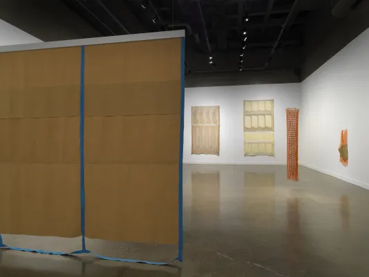 In the foreground on the left, a large brown rectangular tapestry hangs suspended from the ceiling. Blue fiber strips border brown panels with the strips dangling onto the floor. In the background, two large brown and beige tapestries hang on the wall. On the right, two orange netlike sculptures are on display with one hanging off the wall and one suspended from the ceiling.