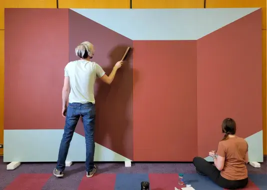 Image of two conservators wearing blue gloves working on repairing a large scale red painting. The person on the left side is standing while the person on the right side is sitting on the floor.