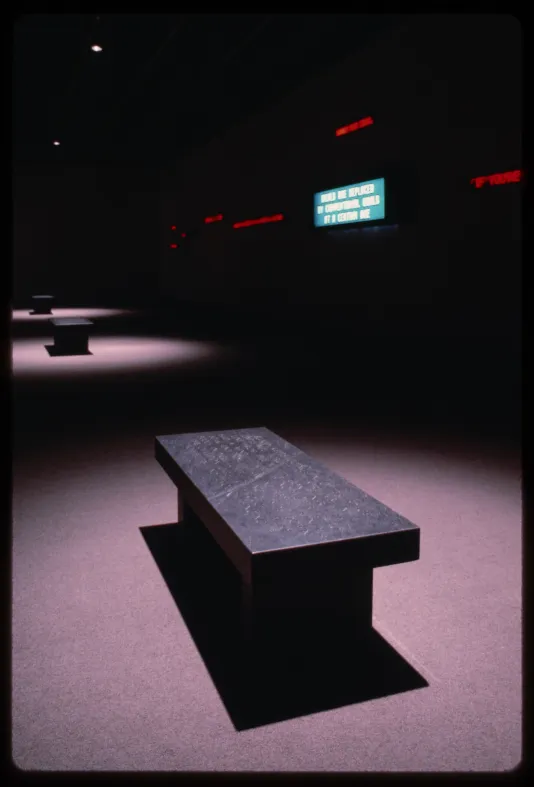 Lit up sign hung on a wall in a dark room with three black benches lit by spotlights.