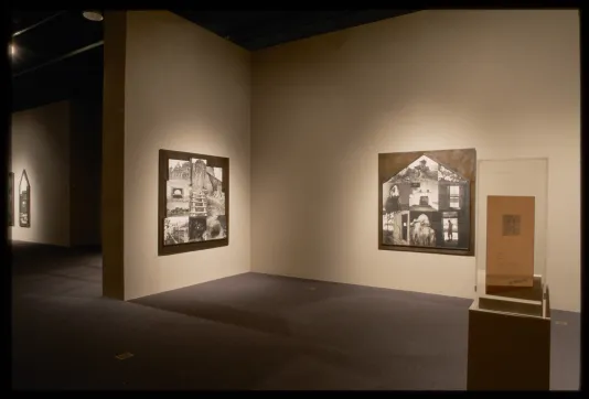 Document sits in glass case in middle of room, with three black and white historical photograph collages hanging on walls.