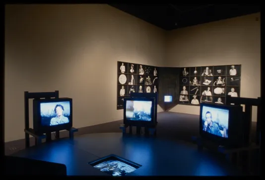 Three tvs with videos of people sit in ring in front of black and white photos of people and items covering the wall corner.