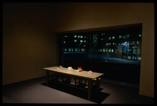 Spotlit table with four books sits in front of large window looking outside to MIT courtyard with lit buildings.