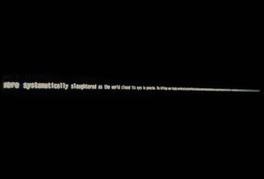 Backlit white words “were systematically slaughtered as the world closed its eyes…” strung across a dark wall.