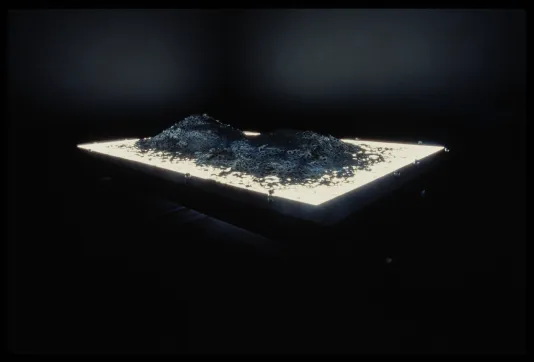 2 mounds of framed photographic slides on a light-table cast a glow in a dark gallery.