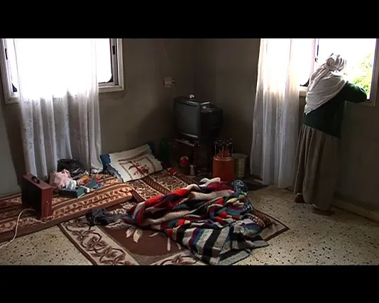 A woman stares out a curtained window, her back to a drab room with concrete colored walls, a futon and bedding on the floor.