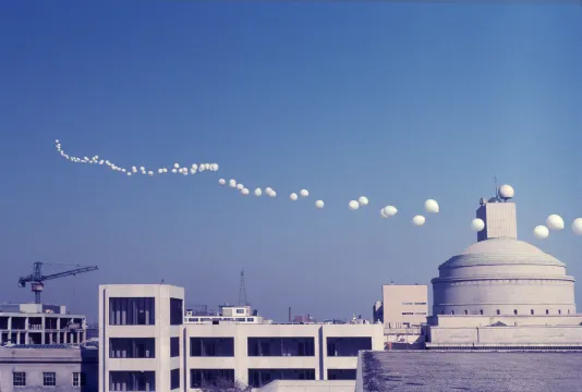 A skyline photo of buildings at MIT, a clear blue sky and a string of large white weather balloons floating into the distance. 