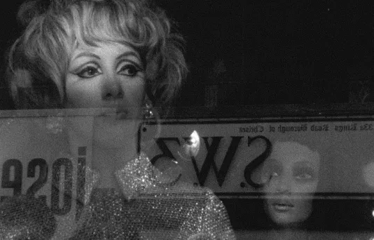 A video still of a woman in a sequined top and eye makeup in a store window with reflections and a female mannequin