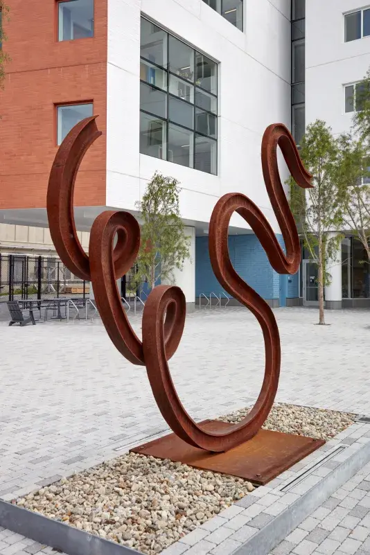 Matt Johnson's Untitled(Swan) sculpture of a warped bent metal train track is pictued in front of MIT campus buildings.